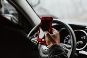person in a car with one hand on the steering wheel and holding a phone in the other hand
