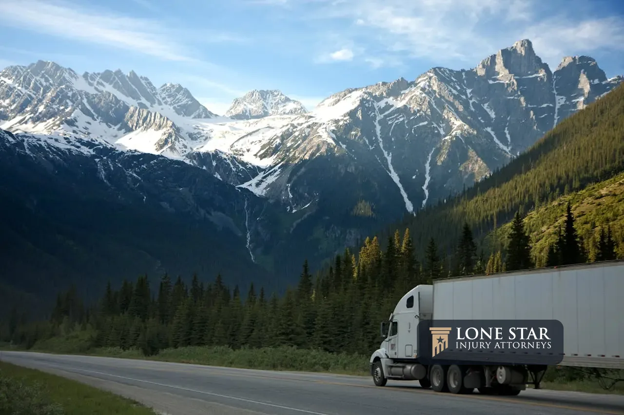 Commercial semi-truck traveling along a mountain highway surrounded by forest and snow-capped peaks.